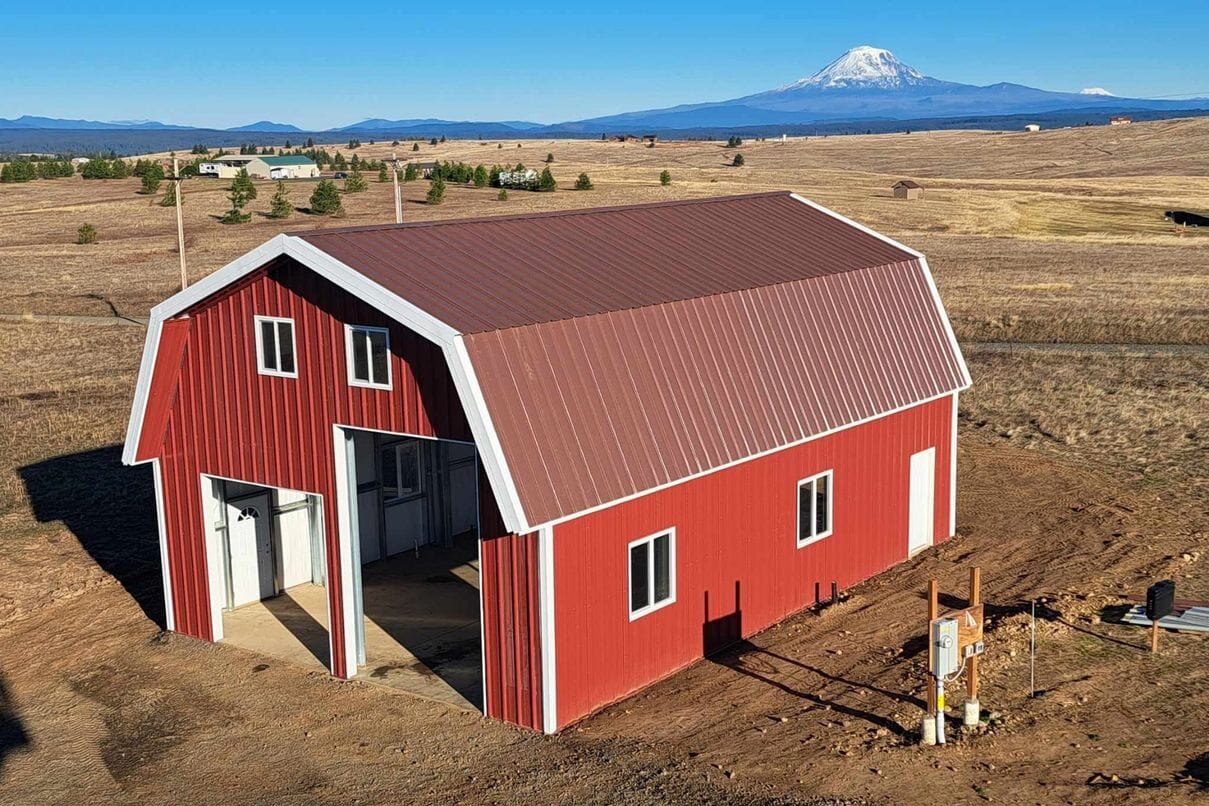 A red steel barn built by Longhorn Structures on open ground near Klamath Falls, with snow-capped mountains behind it